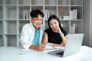 Genç bir çift, modern oturma odalarındaki bir masaya oturmuş, faturaları gözden geçiriyorlar ve birikimlerini dizüstü bilgisayarla planlıyorlar..