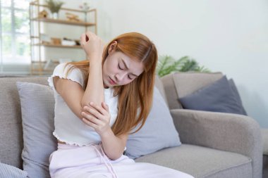 Evdeki kanepede oturan genç bir kadın, dirseğini acıdan tutuyor, rahatsızlık ya da sakatlık belirtileri gösteriyor..