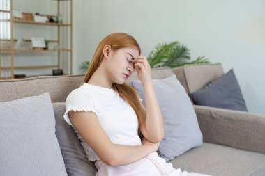 Başı ağrıyan genç bir kadın, modern oturma odasındaki bir kanepede oturuyor, başını acıdan tutuyor..