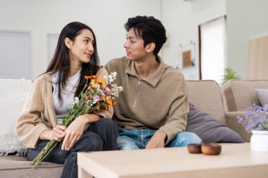 Evdeki kanepede oturan genç bir çift çiçeklerle sevgi dolu bir anı paylaşıyor, modern ve rahat bir yaşam tarzını sergiliyor..