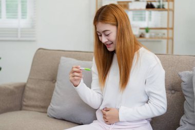 Pozitif bir hamilelik testi tutan neşeli genç bir kadın. Evdeki rahat bir kanepede otururken gülümsüyor ve karnına dokunuyor..