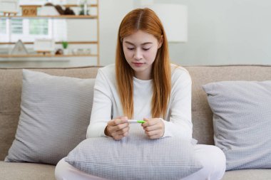 Uzun saçlı genç bir kadın, elinde hamilelik testi olan bir kanepede oturur ve modern bir oturma odasındaki sonuçları yansıtır..