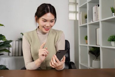 Uzaktan çalışan genç bir kadın, arka planda kitaplıklar ve bitkilerle modern bir ev ofisinde akıllı telefonunu kullanıyor..