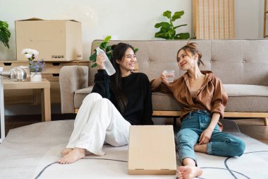 Neşeli lezbiyen bir çift yeni evlerinde eğleniyor, etrafı mobilya ve kutularla çevrili, taşınmalarını içkilerle ve gülümsemeyle kutluyorlar..