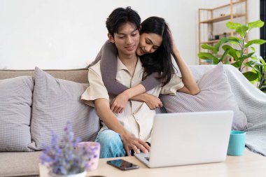 Keyifli bir çift, evde birlikte vakit geçirmenin keyfini çıkarıyor. Kanepede dizüstü bilgisayarlarıyla oturuyorlar. Etrafı rahat ve modern oturma odalarıyla çevrili..