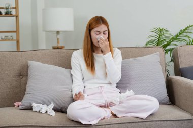 Soğuk algınlığı geçiren genç bir kadın, kanepede oturuyor, burnunu mendile sümkürüyor, buruşuk mendillerle çevrili..