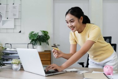 Sarı gömlekli genç bir kadın, modern ofis ortamında bilgisayarında online bankacılık için kredi kartı kullanıyor..