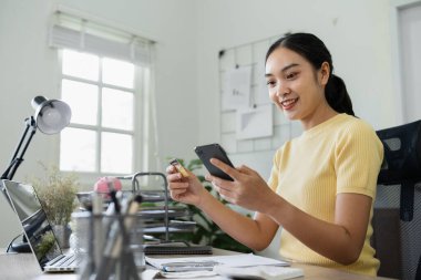 Modern bir ev ofisteki genç bir kadın internet bankacılığı, sergi teknolojisi ve kolaylık için kredi kartını ve akıllı telefonunu kullanıyor..