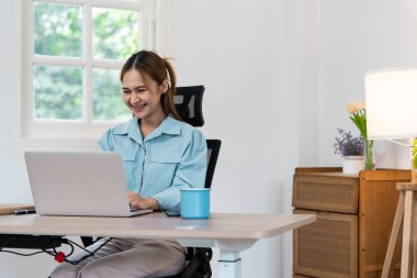 Ev ofisinden uzaktan çalışan, gülümseyen ve dizüstü bilgisayarıyla meşgul genç bir kadın. Doğal ışık ve rahat dekorlu modern çalışma alanı.