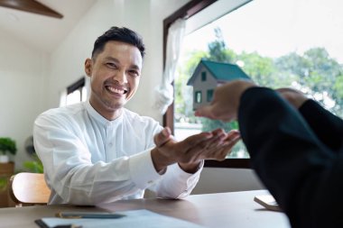 Bir emlakçı, başarılı bir emlak anlaşmasını sembolize ederek parlak bir ofisteki gülümseyen bir müşteriye ev modelini teslim eder..