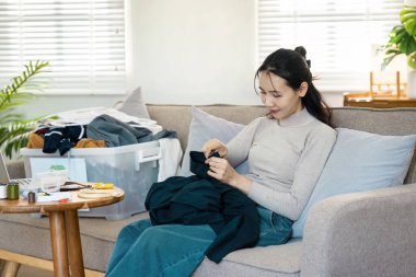 Bir kadın, parlak ve rahat bir oturma odasında dikiş dikerek ve onararak elbiselerini geri dönüşüm için kanepeye oturur..