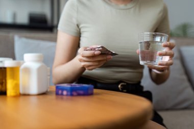 Evdeki ilaçlarını yöneten bir kadın, elinde haplar ve bir bardak suyla, masada çeşitli ilaç şişeleriyle, ev sağlığı ve ilaç yönetimini ön plana çıkarıyor..