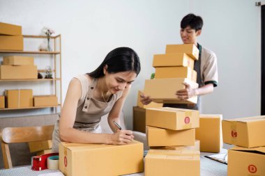 Küçük işletme sahipleri, bir ev ofisinde online siparişler için karton kutuları paketleyip etiketliyor, takım çalışması ve adanmışlık sergiliyorlar..