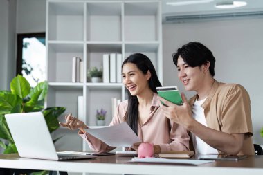 Finansal planlama için tablet kullanan genç bir çift, bir domuz kumbarası, faturalar ve defterlerle çevrili, birikimleri ve bütçeleri sembolize eden.