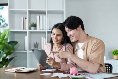 Finansal planlama için tablet kullanan genç bir çift, bir domuz kumbarası, faturalar ve defterlerle çevrili, birikimleri ve bütçeleri sembolize eden.