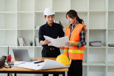 İki mühendis modern bir ofisin planlarını tartışıyorlar, güvenlik donanımı giyiyorlar, inşaat aletleri ve belgelerle çevrili..