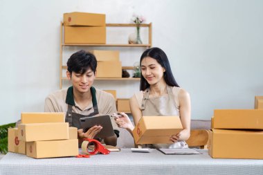 İki genç girişimci e-iş operasyonlarını yönetmek için dijital cihazlar kullanıyor, modern bir çalışma alanındaki paketleme kutularıyla çevrili..