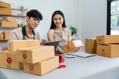 İki genç girişimci, e-iş operasyonlarını yönetmek için dijital cihazlar kullanıyorlar. Etrafları, modern bir çalışma alanındaki paketleme kutularıyla çevrili..