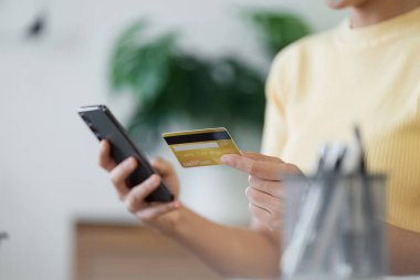 Elinde kredi kartı ve akıllı telefon olan bir kadının, modern bir ev ortamında online alışveriş yaparken yakın plan fotoğrafı..