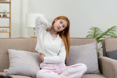 Genç bir kadın kendini iyi hissetmiyor, koltukta oturuyor, boynunu tutuyor ve sırtı ağrıyor, rahat kıyafetler giyiyor..