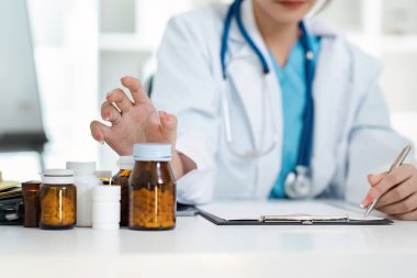 Steteskoplu beyaz önlüklü bir doktor reçete şişelerini inceler ve sağlık hizmetlerini ve tıbbi uygulamaları sembolize eden notlar yazar..