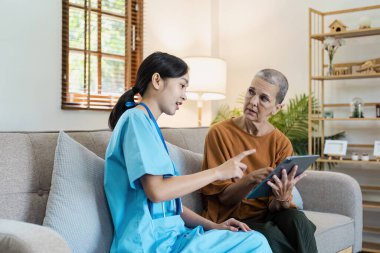 Mavi önlüklü bir sağlık uzmanı, evde yaşlı bir kadına kişisel tıbbi bakım ve destek için dijital tablet kullanarak yardımcı oluyor..
