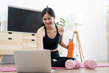 Spor giyim bölümünde yoga yapan genç bir kadın rehberlik için dizüstü bilgisayar kullanıyor. Yanında dambılları olan bir su şişesi tutuyor, sağlıklı ve aktif bir yaşam tarzını teşvik ediyor..