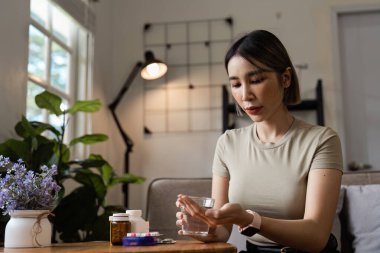 Bir kadın masada oturuyor, ilaç almaya hazırlanıyor, etrafında reçeteli şişeler ve rahat bir ev ortamı var..