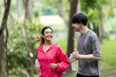 Neşeli bir çift yemyeşil bir parkta birlikte koşuyor, ellerinde su şişeleri ve güneşli bir günün tadını çıkarıyorlar..