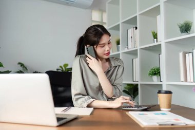 Modern bir ev ofisinde bilgisayar, akıllı telefon ve hesap makinesi kullanarak uzaktan çalışmayı ve üretkenliği temsil eden genç bir kadın..