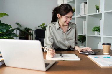 Evinden çalışan genç bir kadın, bilgisayar ve hesap makinesi kullanarak çevresi bitki ve ofis malzemeleriyle çevrili, modern bir ofis masasında çalışıyor..
