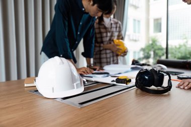 Modern bir ofiste planlar ve inşaat planlarını tartışan mimar. Masanın üzerinde şapka ve kulak koruyucusu da dahil olmak üzere güvenlik teçhizatı var..