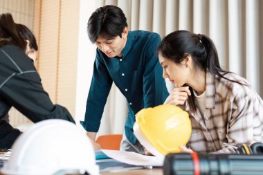 Modern bir ofis ortamında inşaat planlarını planlar ve güvenlik teçhizatlarıyla tartışan bir grup genç profesyonel..