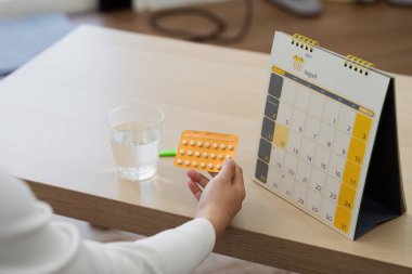Doğum kontrol haplarını elinde takvim ve bir bardak suyla ahşap bir masada tutan bir kadının yakın plan doğum kontrolü ve aile planlamasını sembolize ediyor..