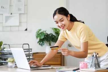 Bilgisayarında online alışveriş yapmak için kredi kartı kullanan gülümseyen genç bir kadın rahat bir ofis ortamında..