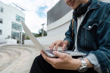 Kampüste dizüstü bilgisayar kullanan, gülümseyen ve kulaklık takan bir üniversite öğrencisi..