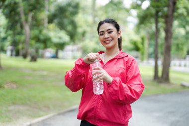 Kırmızı ceketli, elinde su şişesi olan gülümseyen genç bir kadın yemyeşil bir parkta spor yaparken mola veriyor..
