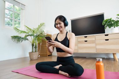 Aktif giyimli genç bir kadın evde sağIık pratiği yapıyor yoga minderinde oturuyor, kulaklıkla müzik dinliyor ve akıllı telefon kullanıyor..