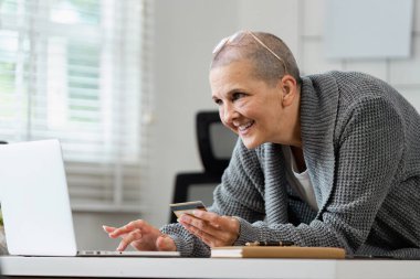 Rahat bir ofis ortamında çevrimiçi alışveriş için kredi kartı ve dizüstü bilgisayar kullanırken gülümseyen yaşlı kadın.