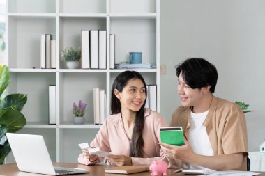 Genç bir çift, tasarruf ve finansal planlarını tartışırken, modern bir ofis ortamında ellerinde bir kumbara ve belgeler tutuyorlardı..