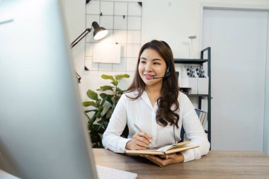 Neşeli bir çağrı merkezi ajanı, kulaklık takıyor. Çağdaş bir ofis ortamında masasında çalışırken not alıyor..