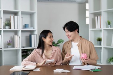 Genç bir çift, tasarruf ve finansal planlarını tartışırken, modern bir ofis ortamında ellerinde bir kumbara ve belgeler tutuyorlardı..