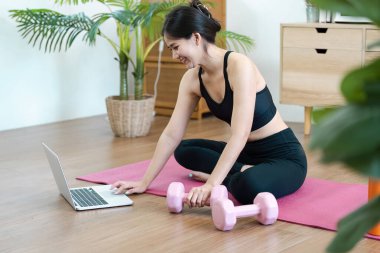 Aktif giyimli genç bir kadın evde yoga yapıyor. Rehberlik için dizüstü bilgisayar kullanıyor. Etrafı bitkiler ve sağlık ekipmanlarıyla çevrili..