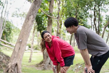 Yeşil bir parkta egzersiz rutinine ara veren genç bir çift, gülümsüyor ve su şişelerini tutuyor, açık havanın tadını çıkarıyor..