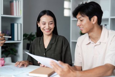 Gülümseyen genç çift finansal belgeleri inceliyor ve modern bir ofis ortamında tasarruf stratejilerini tartışıyorlar masada bir kumbara ile.