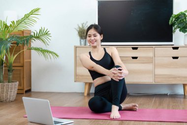 Atletik bir kadın modern bir oturma odasında pembe bir paspasın üzerinde yoga yapıyor, rehberlik için dizüstü bilgisayar kullanıyor, sağlığını ve sağlıklı bir yaşam tarzını destekliyor..