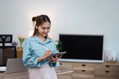 Evden çalışan genç bir kadın, modern ev ofis ortamında dijital tablet kullanıyor, uzak çalışma yaşam tarzını gösteriyor..