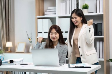 Modern bir ofiste iki iş kadını, belgeler ve ofis malzemeleriyle çevrili bir dizüstü bilgisayarla çalışırken başarıyı kutluyorlar..