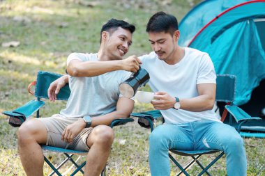İki arkadaş kamp sandalyelerinde oturur, kahve içerler ve çadırın yanında huzurlu bir sabah geçirirler..