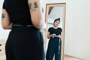 Şişman bir kadın, aynanın önünde belini ölçüyor, vücudun pozitifliğini, kendini kabulünü ve sağlıklı yaşam tarzını destekliyor..